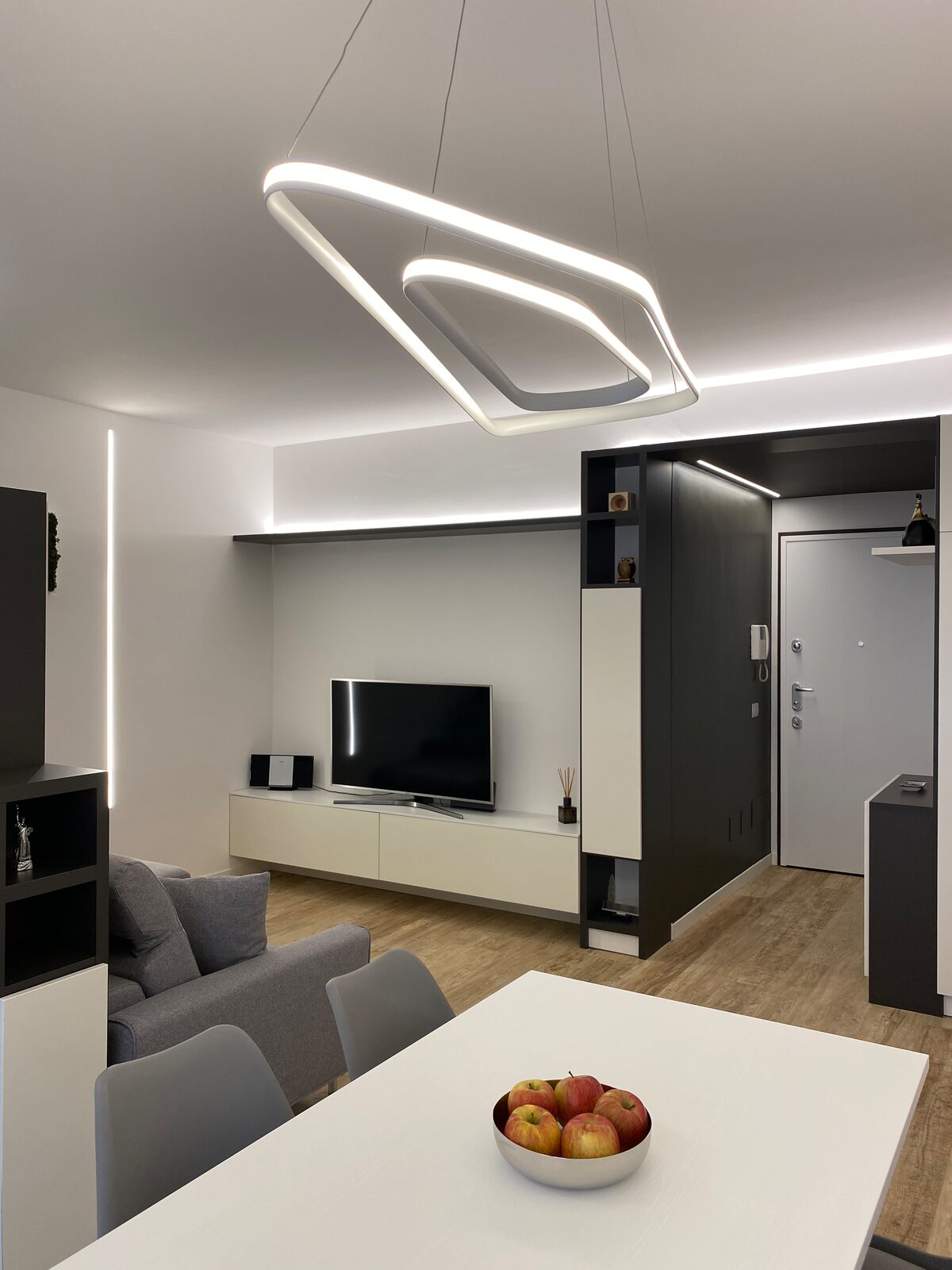Un soggiorno illuminato da linee di luce lungo le pareti e la mensola, con una lampada a sospensione bianca a due anelli sopra il tavolo e l'ingresso in un blocco grigio scuro