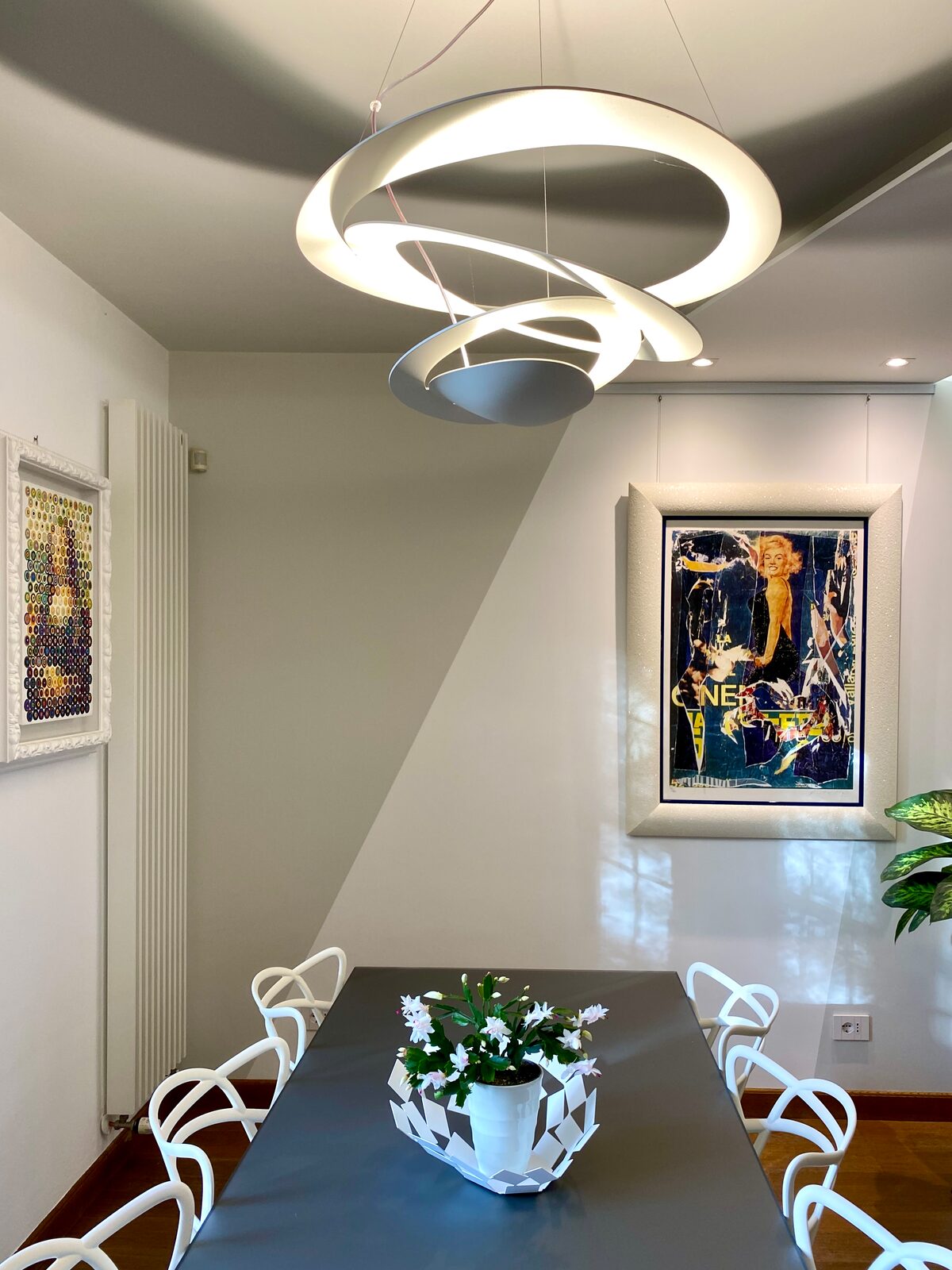 Una sala da pranzo con una lampada a sospensione bianca a spirale sopra il tavolo scuro, sedie bianche, due quadri e la luce del giorno sulle pareti