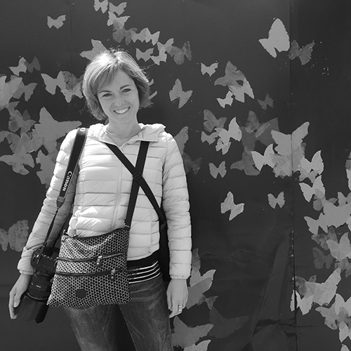 Laura smiles in front of a wall of paper butterflies, with a bag across her chest