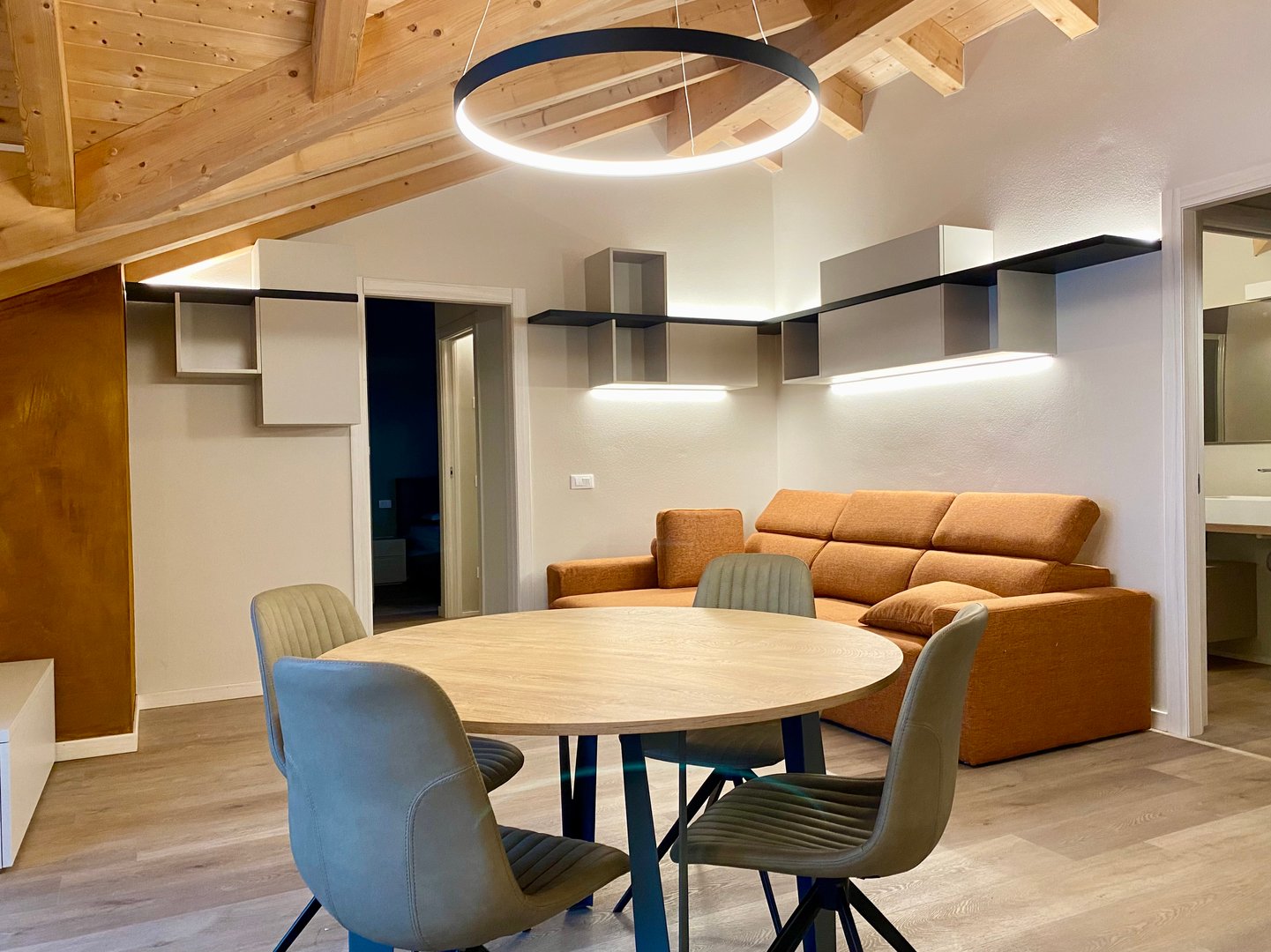 An attic living room with exposed wooden beams, a glowing ring lamp over the round table, lit shelves and an orange sofa