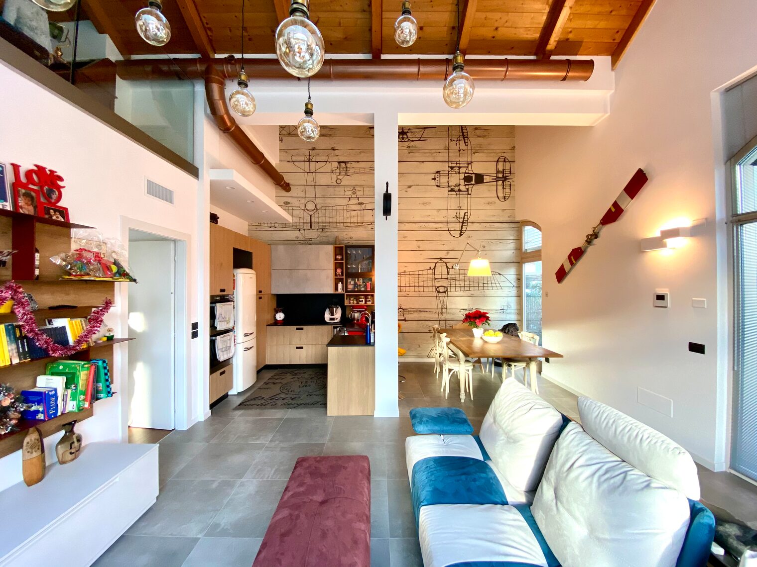 An open space under the wooden beams: the sofa, the kitchen and the dining table in front of a plank wall with drawings of old airplanes, beneath bare-bulb pendant lamps