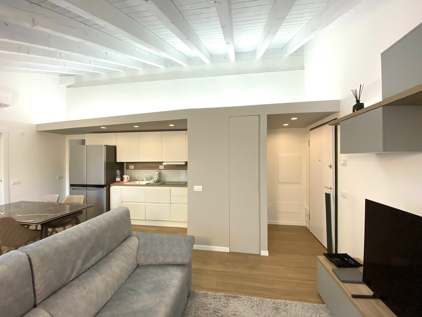 A living room with white wooden beams, a gray sofa, and the kitchen and entrance in the background