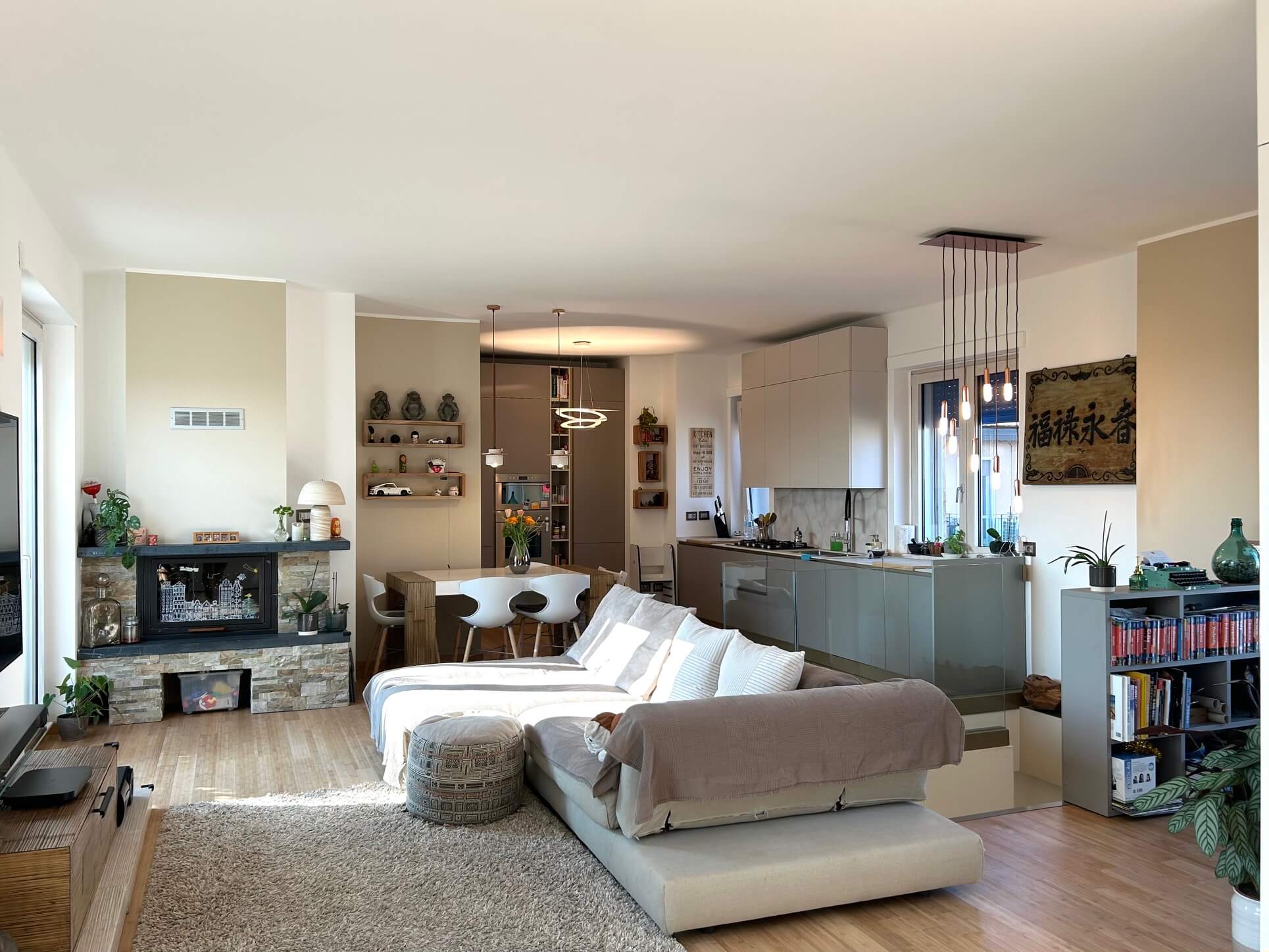 A bright living room with a light sofa, a stone fireplace, a dining table and an open kitchen with a pendant lamp