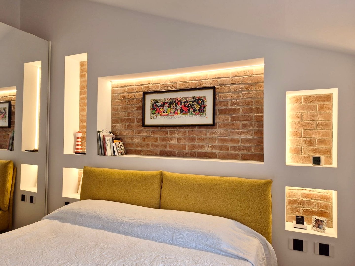 A bedroom with a mustard-yellow headboard and exposed-brick niches lit by strips of light