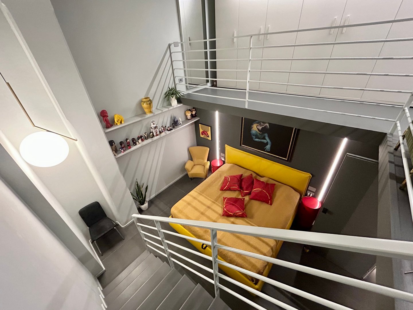 A bedroom seen from the top of the staircase, with the yellow bed under a mezzanine with a white railing