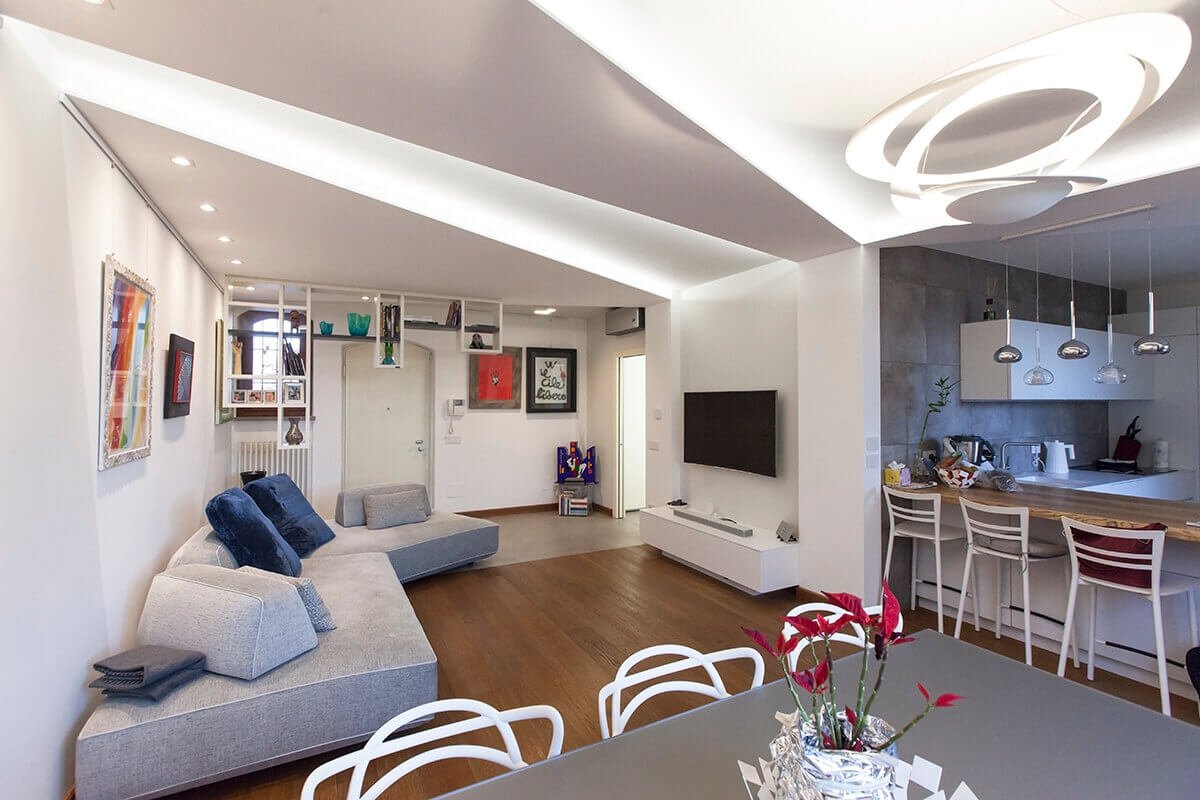 A living room with a continuous-light plasterboard ceiling, a gray sofa and an open kitchen, in a villa